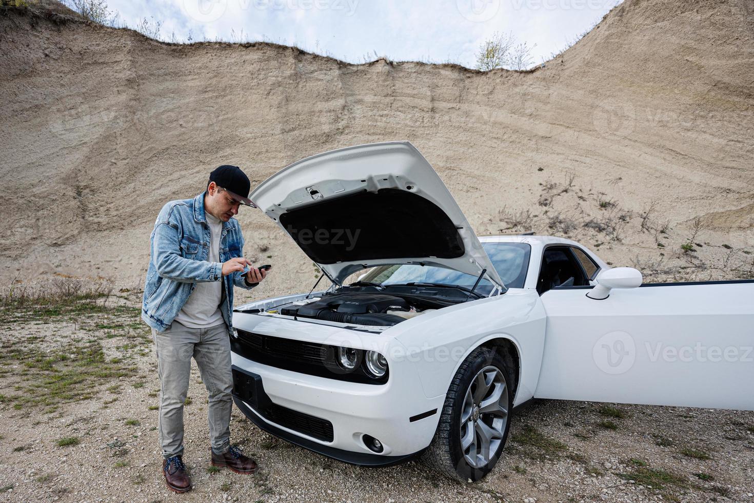 Shocked man is standing near his broken muscle car, open hood, breakdown, need help. Calling to service. photo