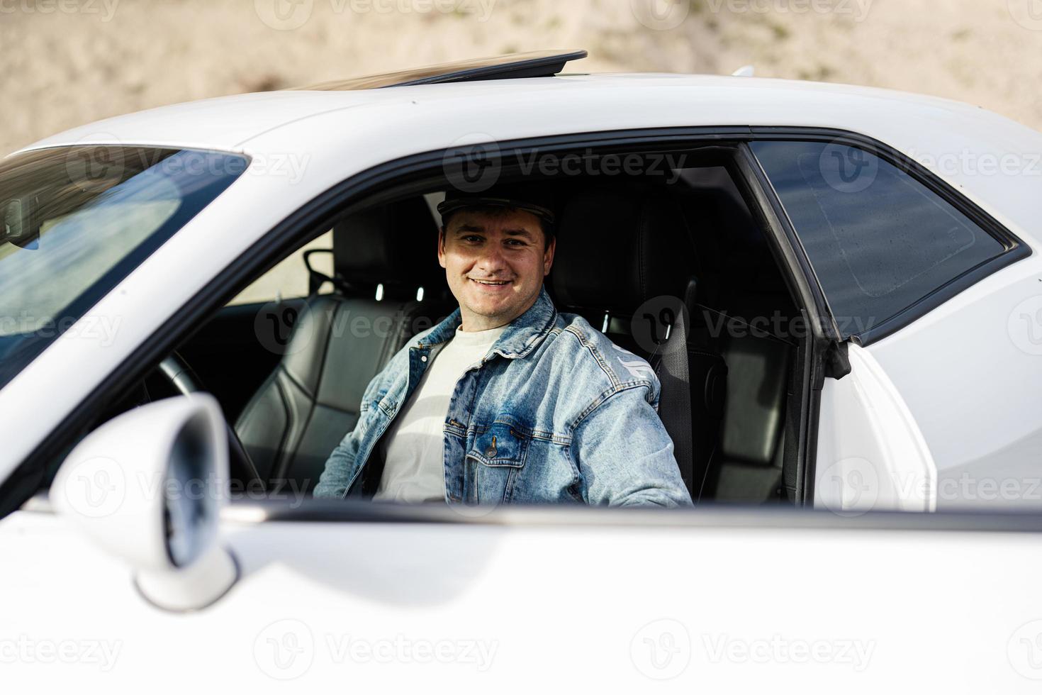 un hombre guapo con chaqueta de jeans y gorra se sienta en su auto blanco. foto