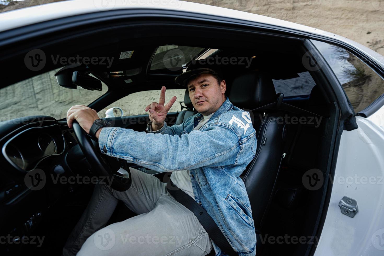 Handsome man in jeans jacket and cap sit at his white muscle car and show two fingers. photo