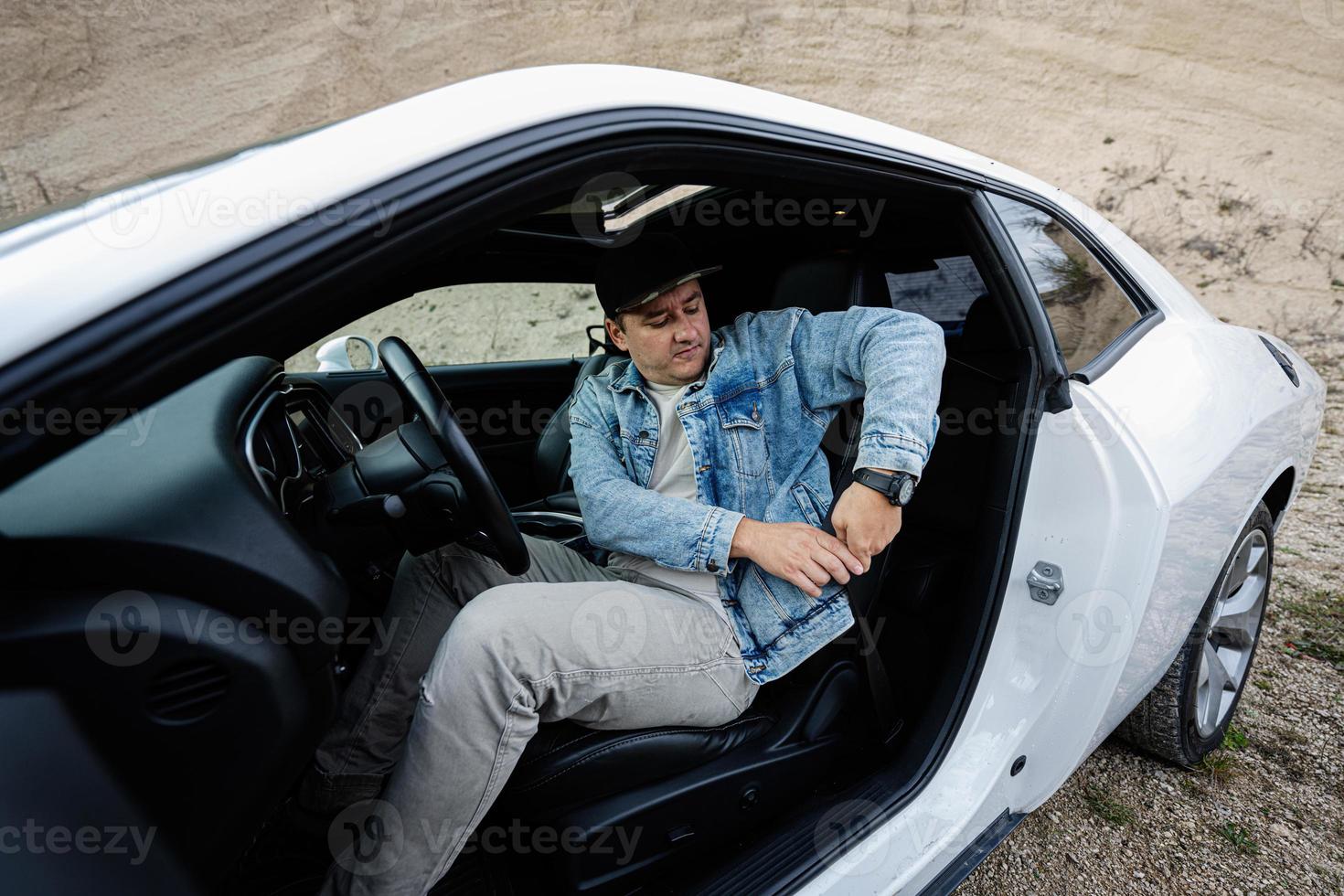 un hombre guapo con chaqueta de jeans y gorra se sienta y ata el cinturón en su auto deportivo blanco. foto