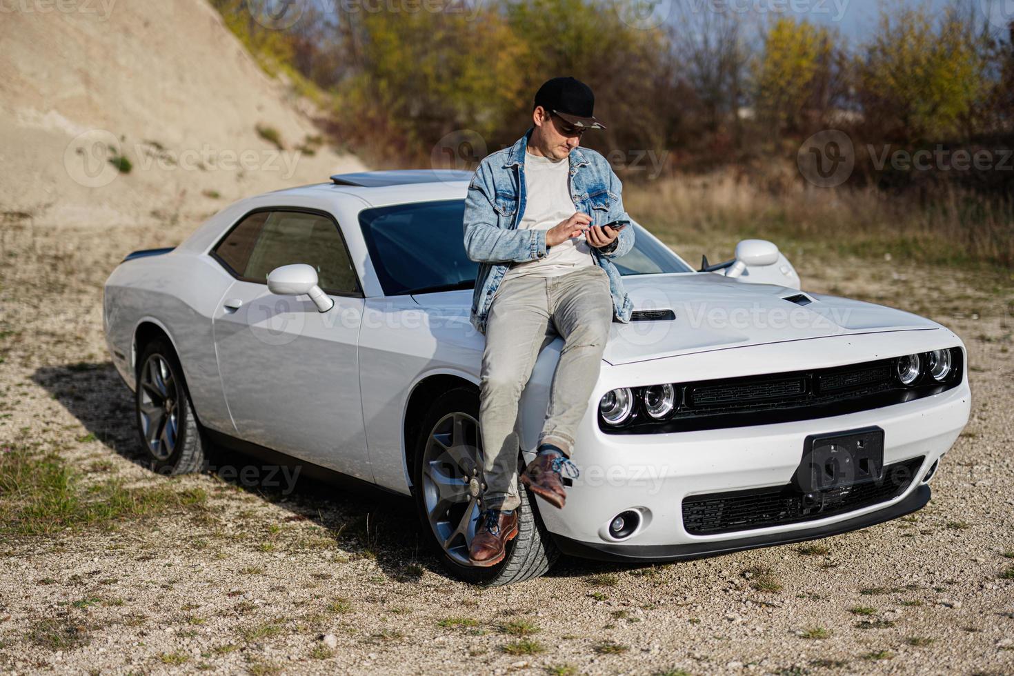 Handsome man in jeans jacket and cap sit on hood at his white muscle car in career and texting on phone. photo