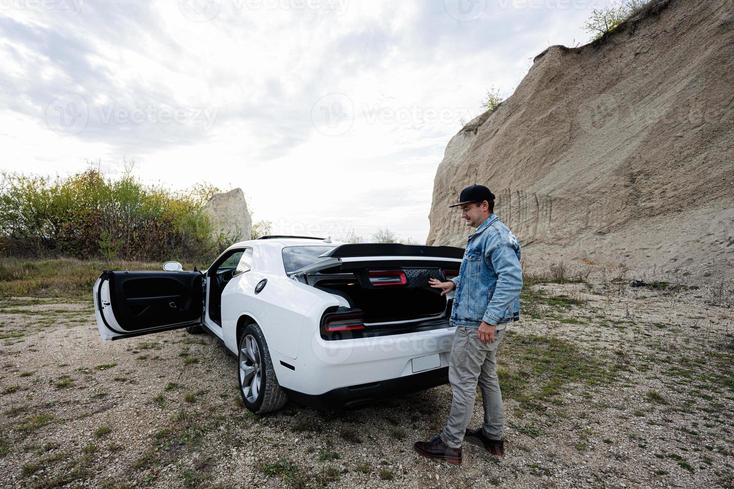 Handsome man in jeans jacket and cap open trunk from his white muscle car in career. photo