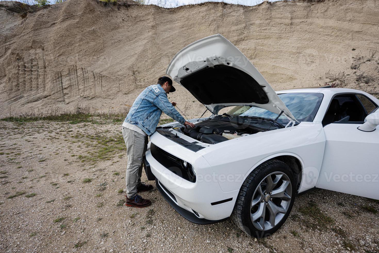 un hombre guapo con chaqueta de jeans y gorra está parado cerca de su auto blanco con capucha abierta, revisa el nivel de aceite del motor. foto
