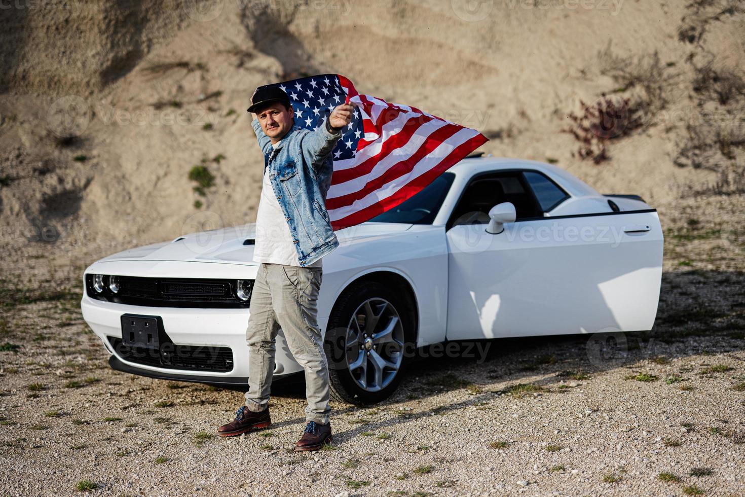 Handsome man in jeans jacket and cap with USA flag near his white american muscle car in career. photo