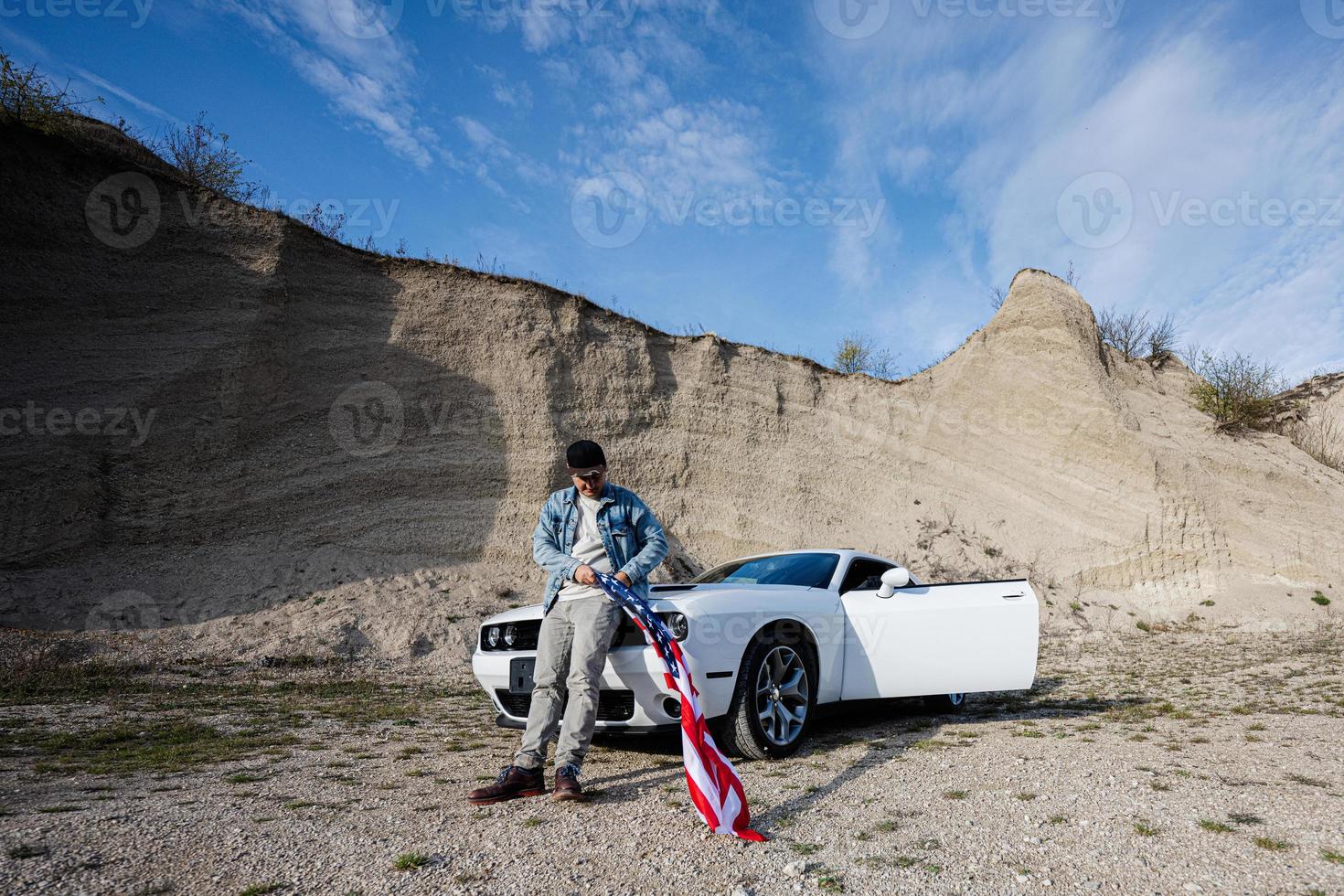 hombre guapo con chaqueta de jeans y gorra con bandera de estados unidos cerca de su muscle car blanco americano en carrera. foto