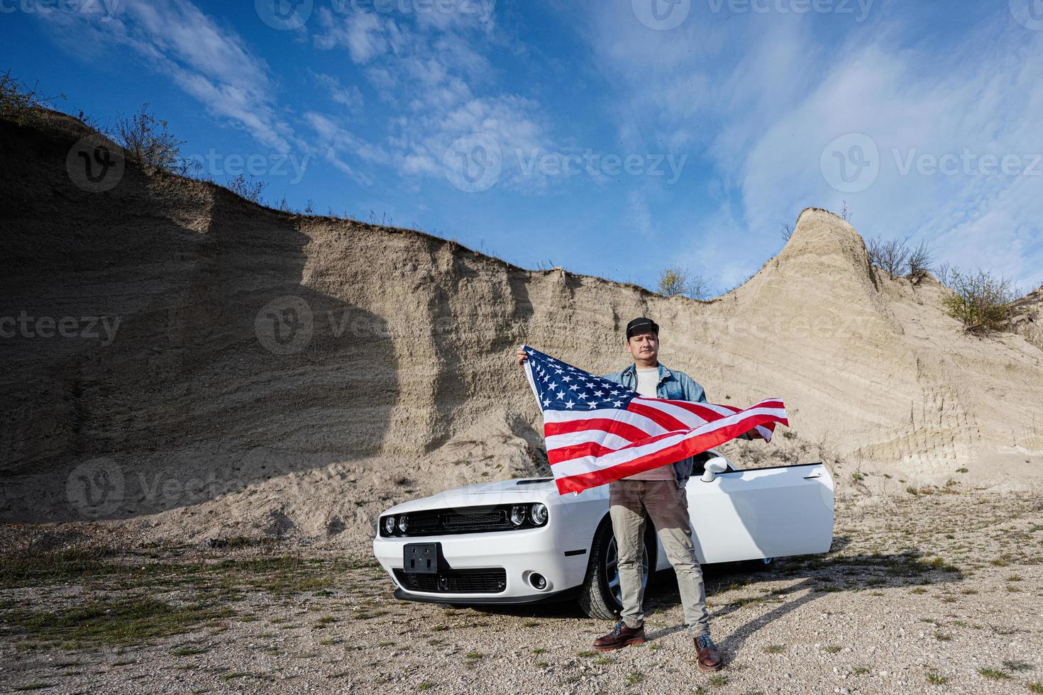 hombre guapo con chaqueta de jeans y gorra con bandera de estados unidos cerca de su muscle car blanco americano en carrera. foto