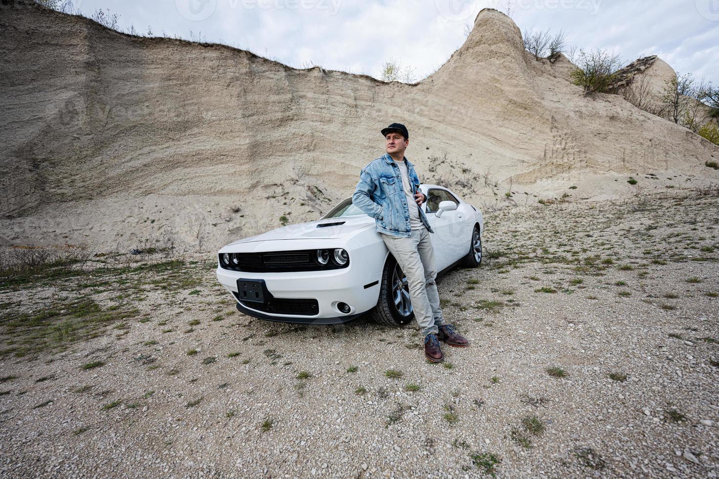 un hombre guapo con chaqueta de jeans y gorra está parado cerca de su auto blanco en carrera. foto