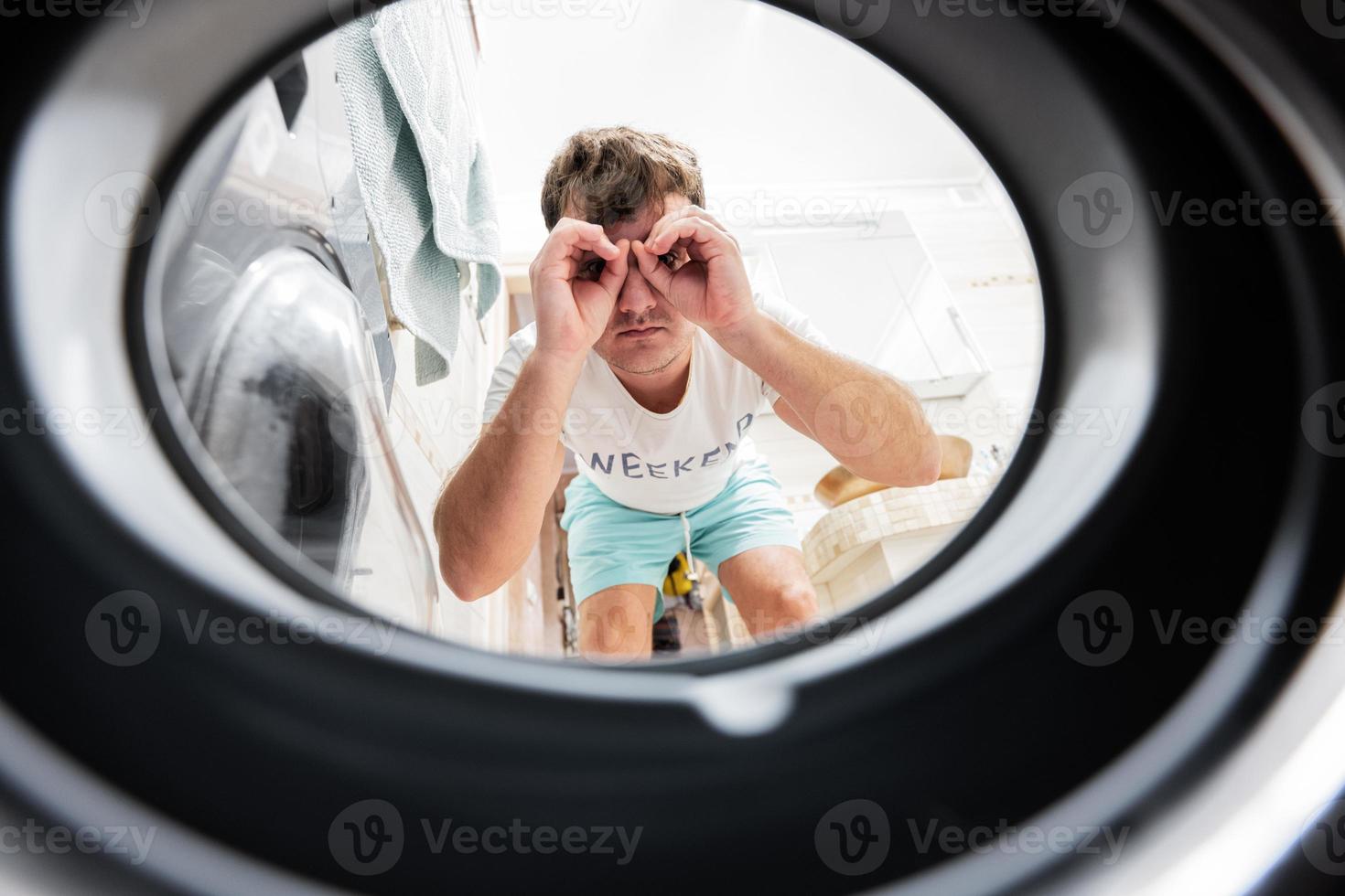 Man view from washing machine inside. Male does laundry daily routine. Looking through binoculars gesture. photo