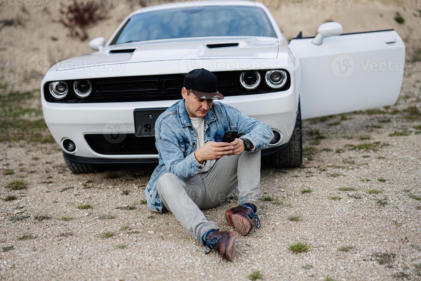 hermoso hombre en pantalones chaqueta y gorra sentado cerca su blanco músculo coche en carrera y mensajes de texto en teléfono. foto