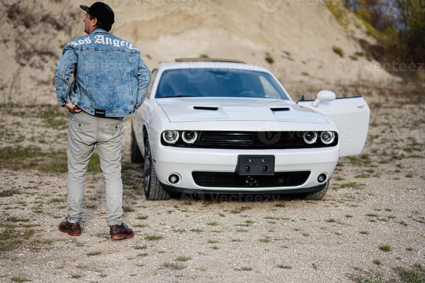 Back of man in jeans jacket and cap near his white muscle car in career. Los Angeles. photo