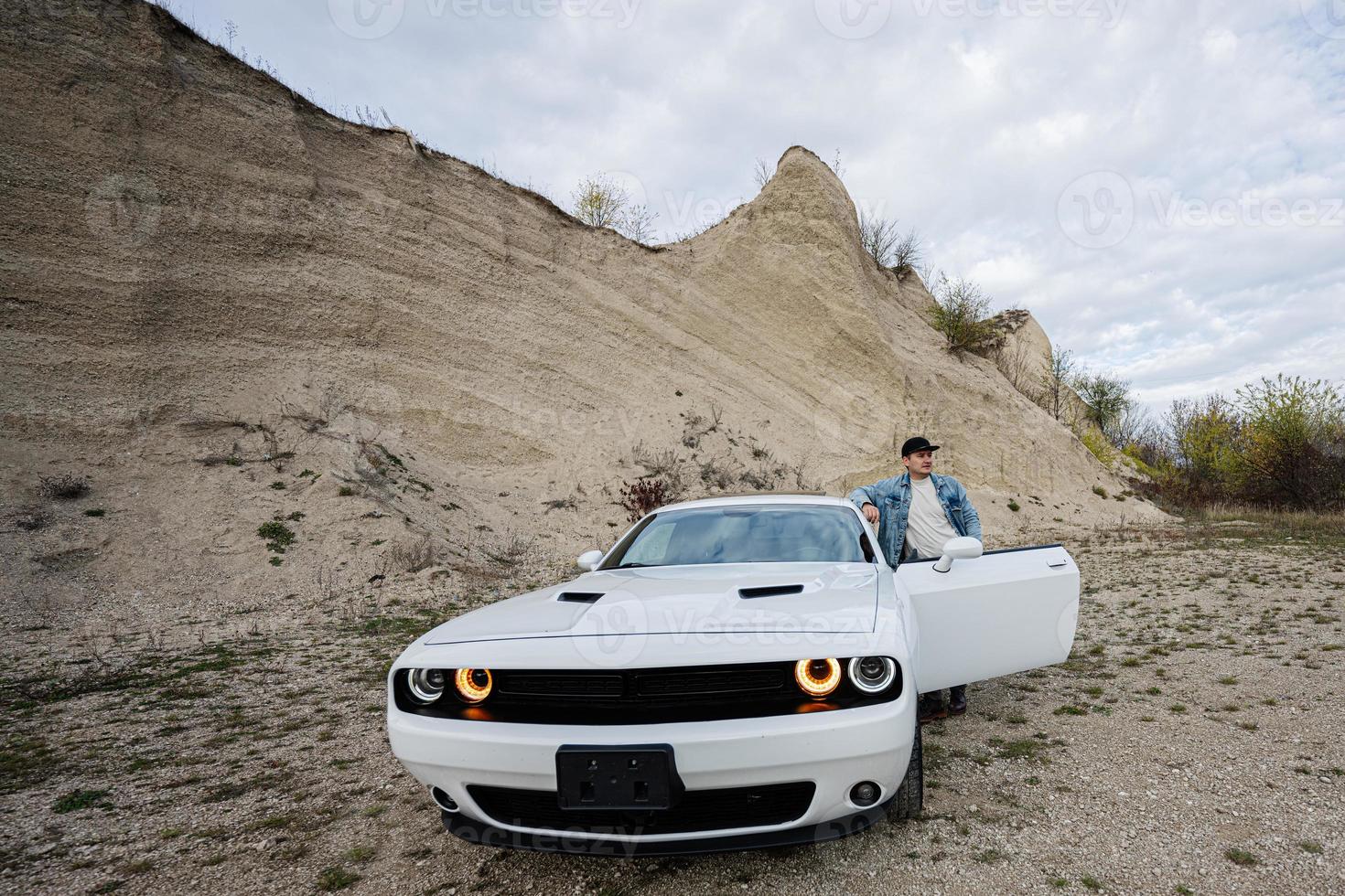 un hombre guapo con chaqueta de jeans y gorra está parado cerca de su auto blanco en carrera. foto