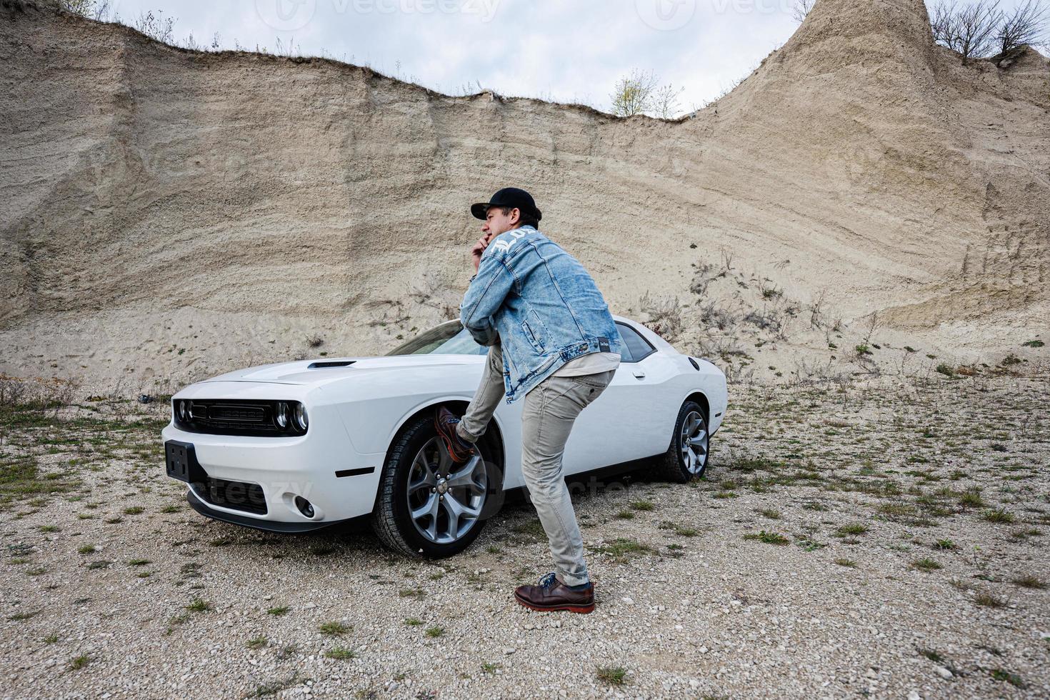 Handsome man in jeans jacket and cap is standing near his white muscle car in career. photo