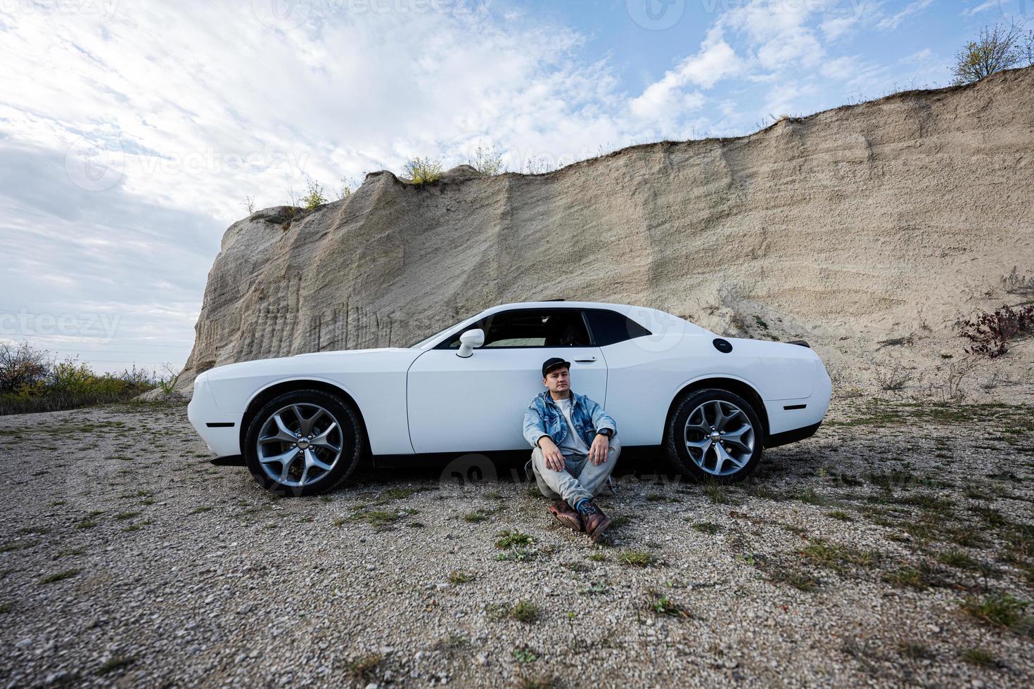 hombre guapo con chaqueta de jeans y gorra sentado cerca de su auto blanco en carrera. foto