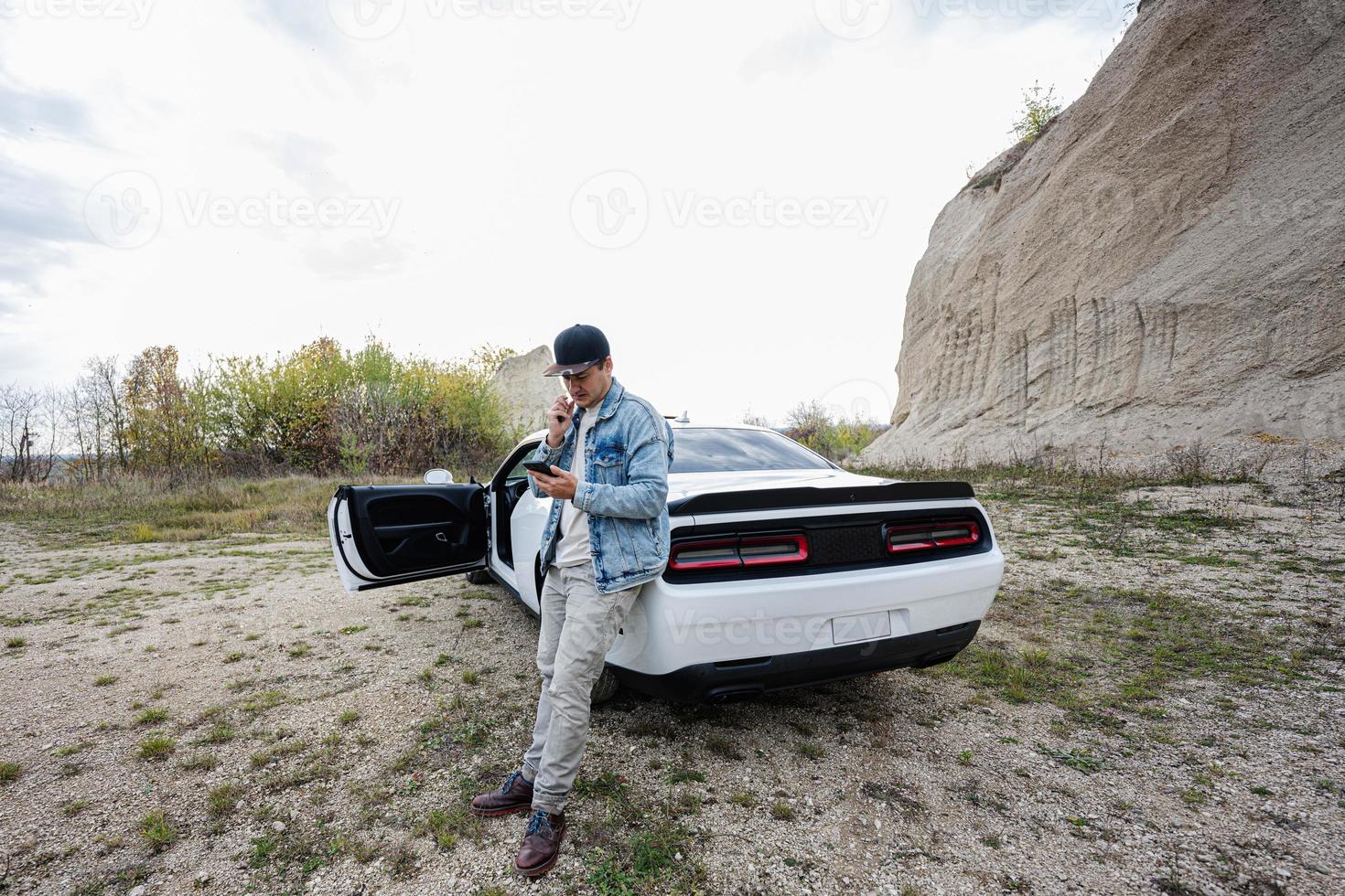 Handsome man in jeans jacket and cap, with mobile phone is standing near his white muscle car in career. photo