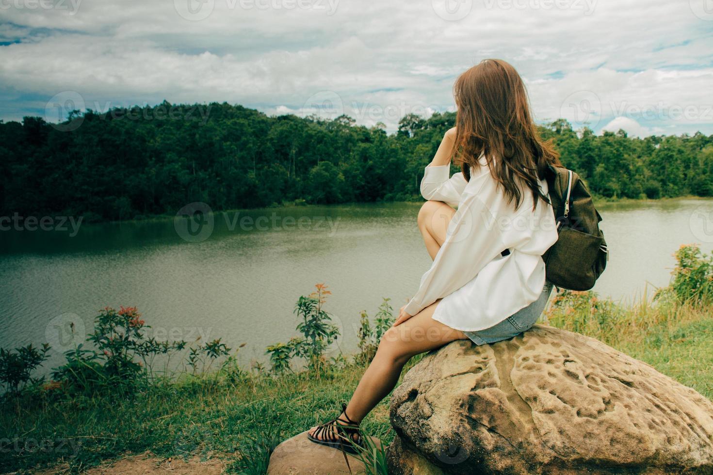 Young asian lonely woman while sitting at stone next to the river among mountains in summer. She was wearing a white shirt shoulder bag, thinking about her problems and felt very sad. photo