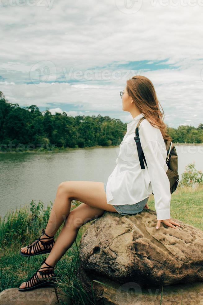 hermosa asiático mujer vistiendo lentes y mochila sentado a Roca siguiente a el río entre montañas en verano. ella estaba vistiendo un blanco camisa hombro bolsa, el clima estaba excelente, sensación relajado. foto