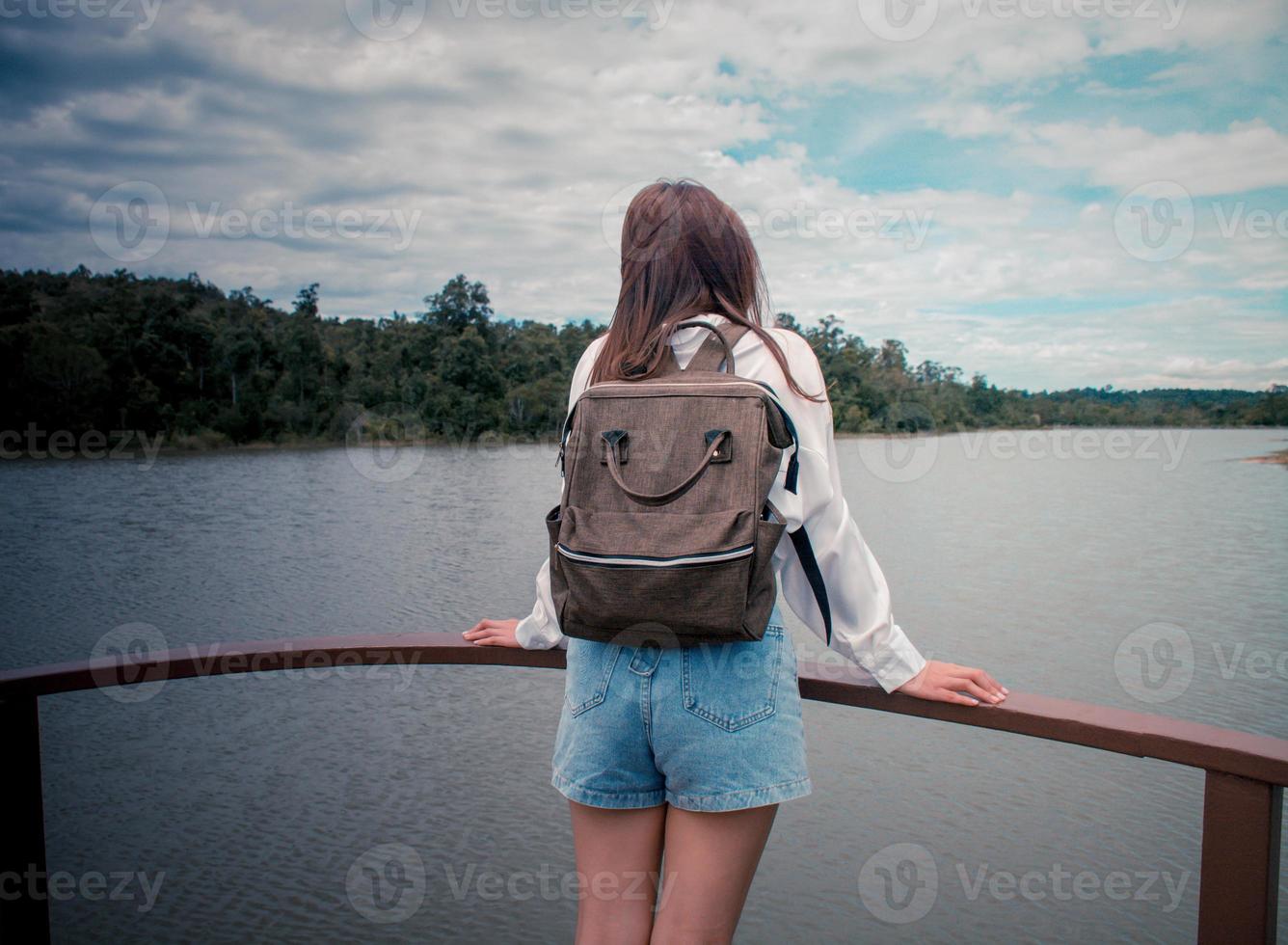 Back view. Young asian lonely woman backpack and standing on the bridge watching the river among mountains in summer. She was wearing a white shirt, thinking about her problems and felt very sad. photo