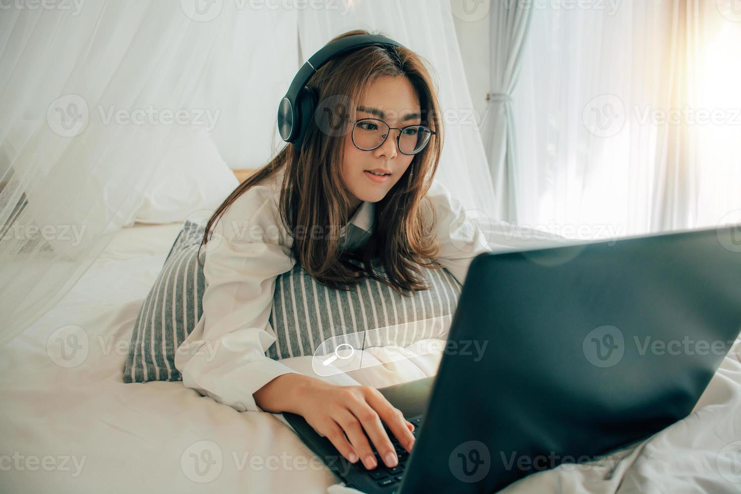 Beautiful young girl wearing glasses and headphones searching Internet Data Information with blank search bar with laptop on the white bed. Business women working with computer notebook finding data. photo