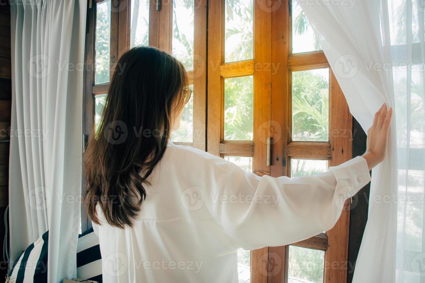 Side view of young woman wearing glasses wake up in bedroom at home or hotel, open white curtains look in window admiring to get fresh air. welcome new day morning feel positive and optimistic. photo