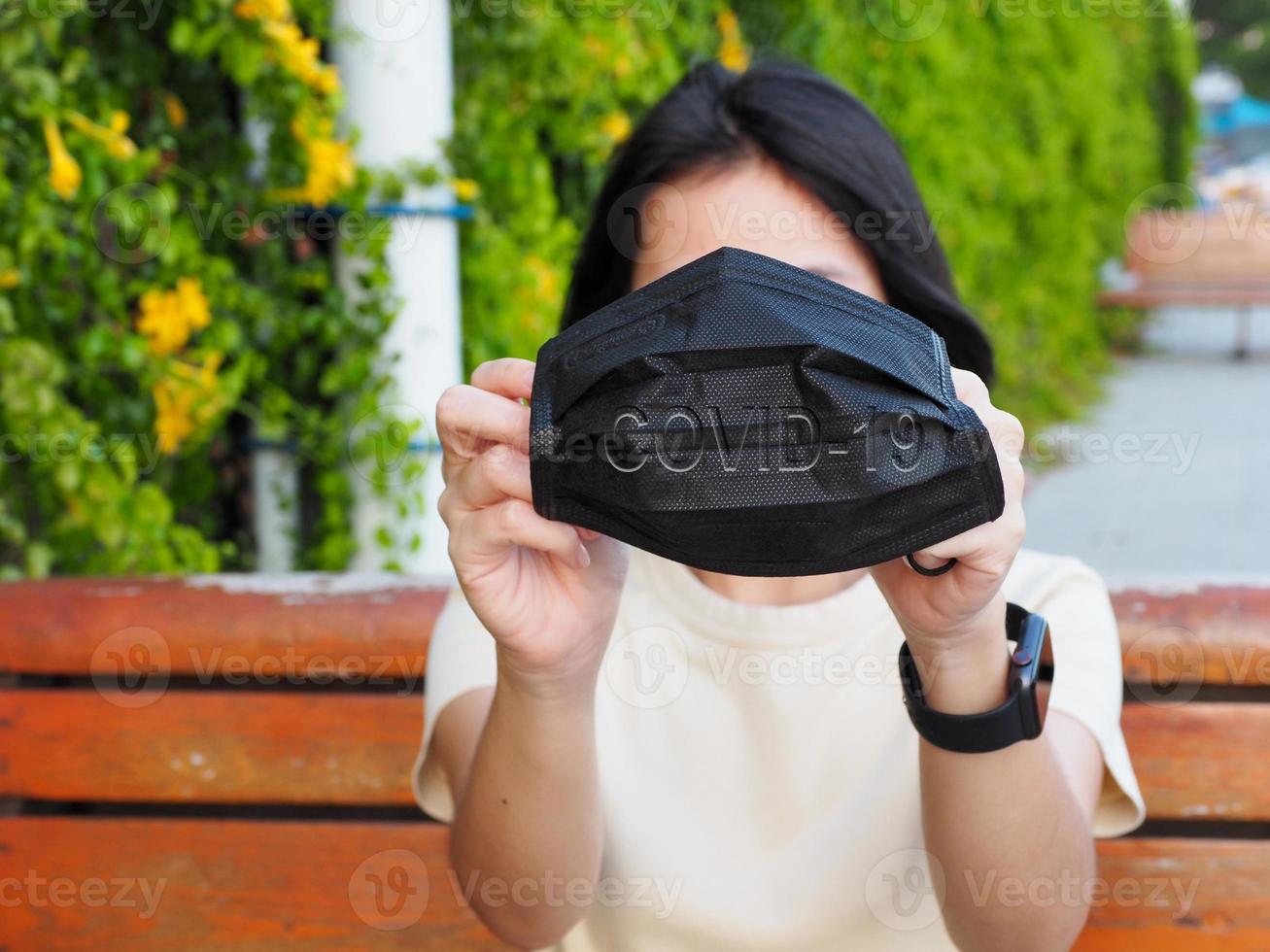 COVID-19 Contagious Hand of a young woman holding a black mask with COVID-19 message in the back inside a park photo