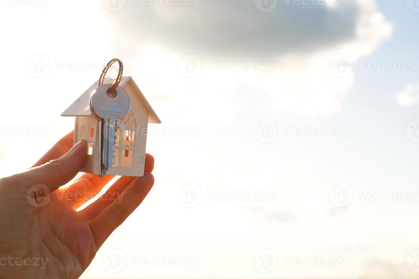 White wooden figure of a house and keys in hand against the background of the sky and field. Dream of your home, building a cottage in the countryside, plan and design, moving to a new house. photo