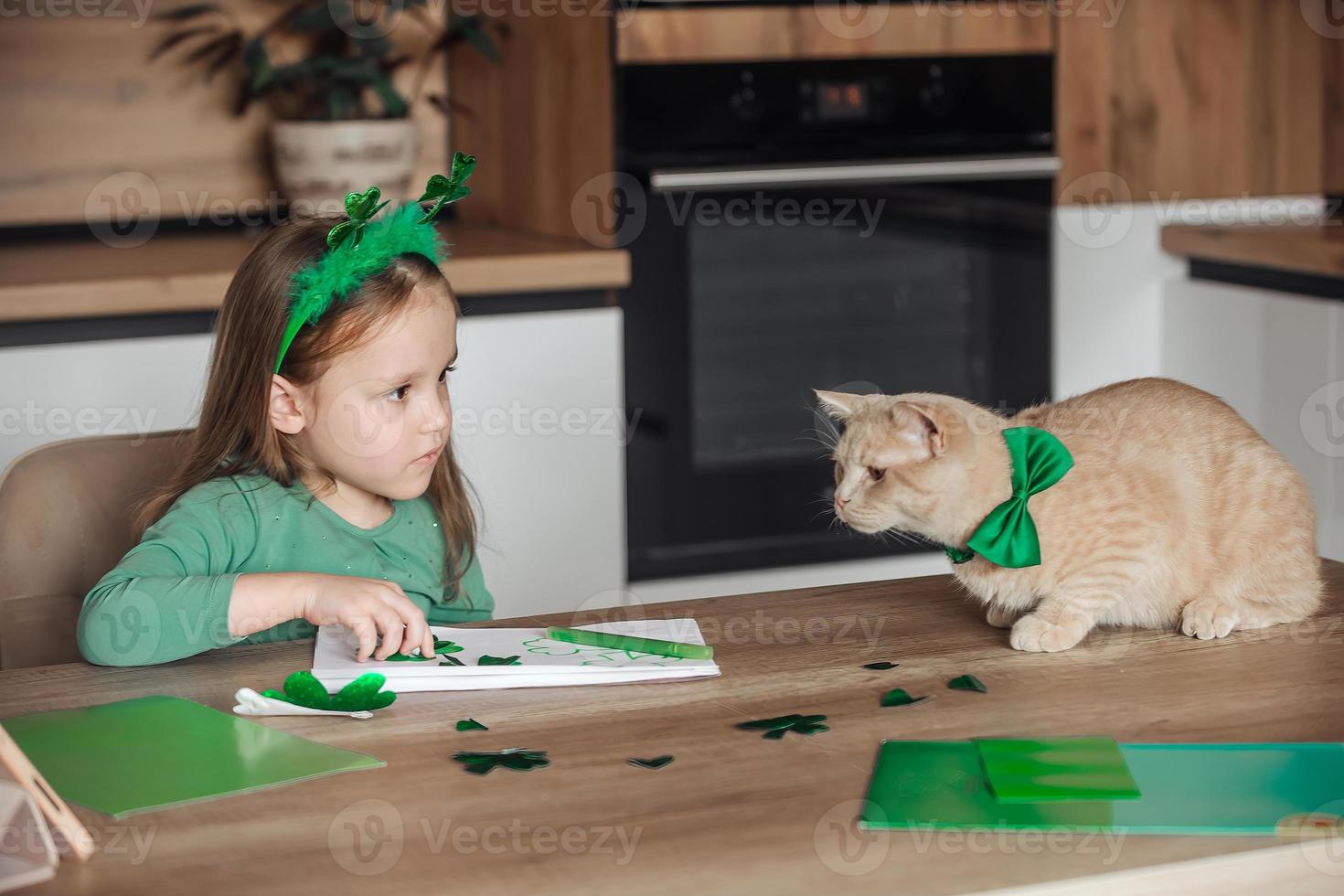 un pequeño niña con un vendaje en su cabeza sorteos y cortes verde tréboles para S t. patrick's día a un mesa a hogar en el cocina, siguiente a su es su hermosa gato con un verde arco Corbata alrededor su cuello foto