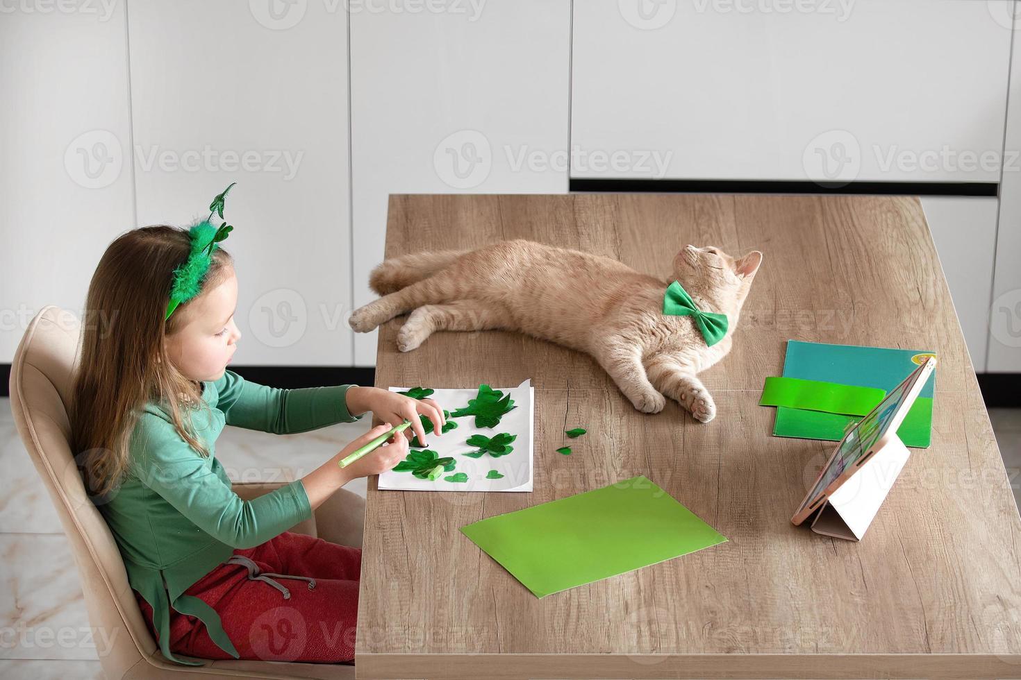 A little girl with a bandage on her head draws and cuts green shamrocks for St. Patrick's Day at a table at home in the kitchen, next to her is her beautiful cat with a green bow tie around his neck photo