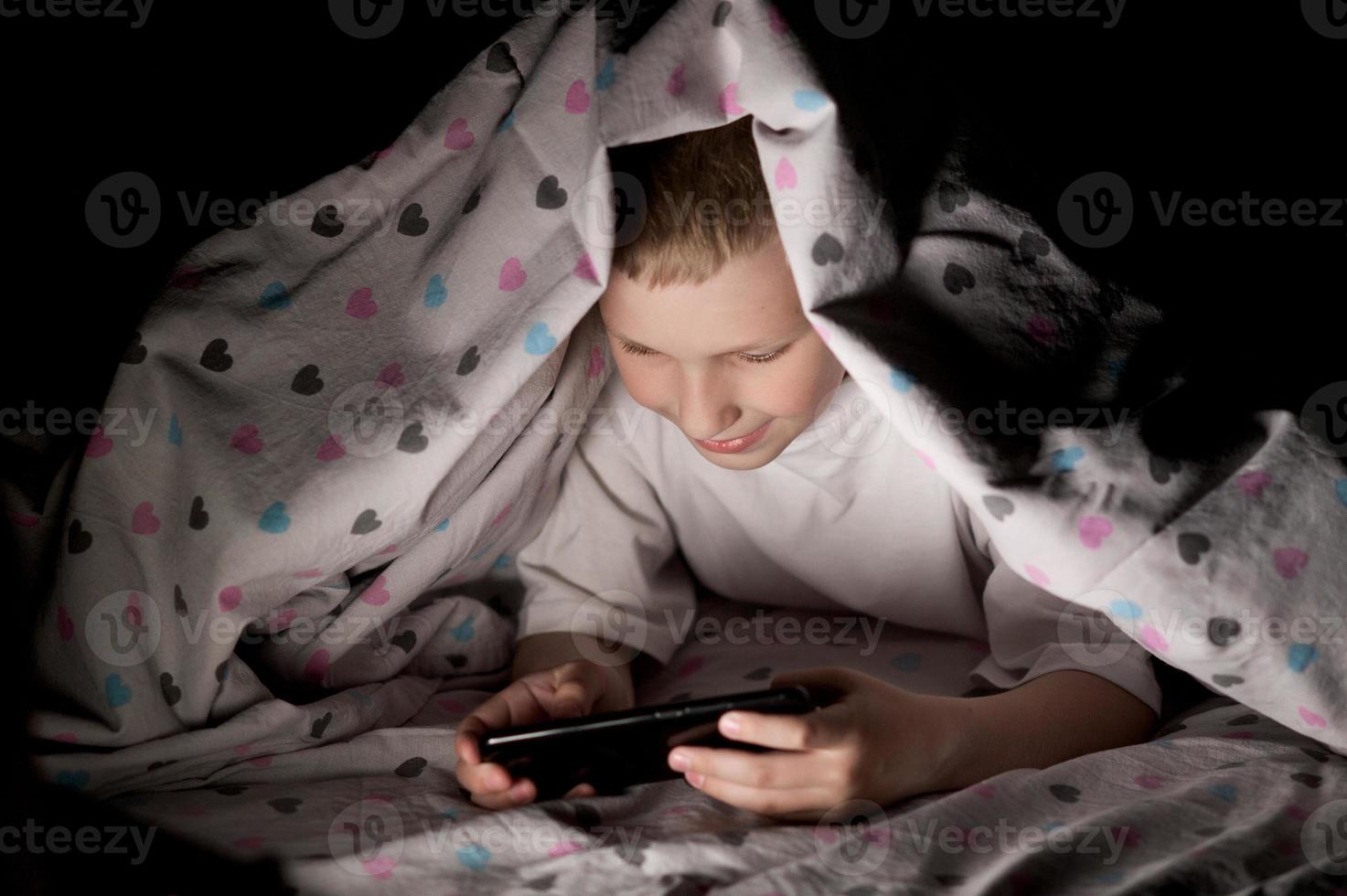 A little boy lies under a blanket at night playing on the phone photo