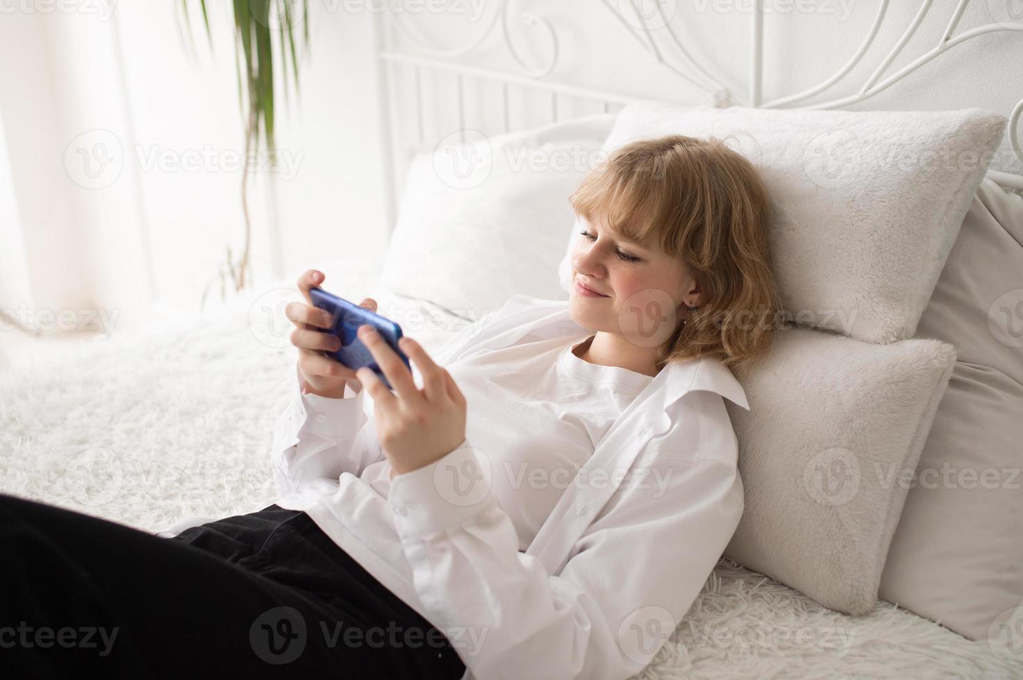 A cute girl in a white shirt and with a phone is lying on the bed. White interior photo