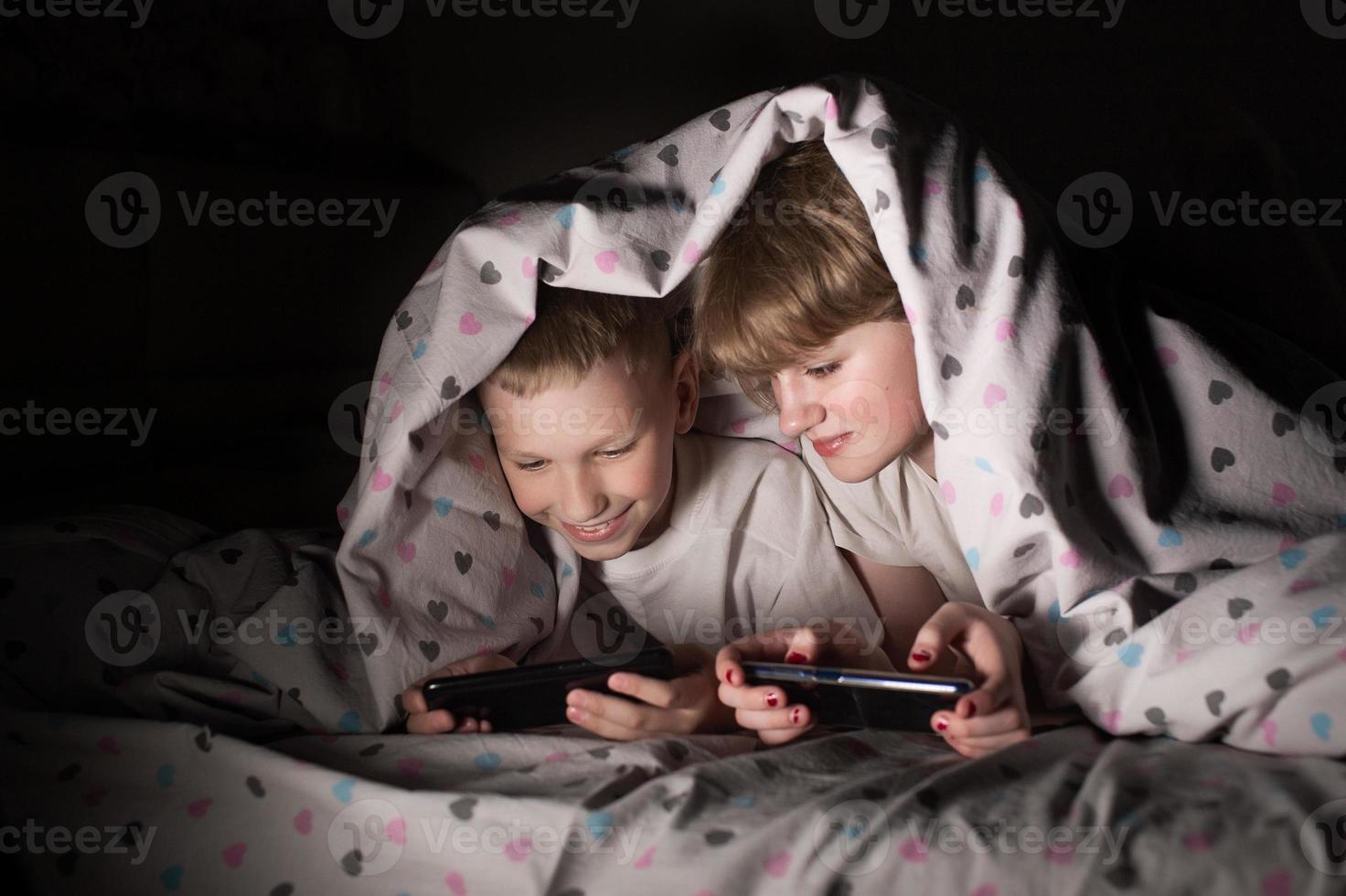 Brother and sister lie under a blanket at night and play on the phone photo