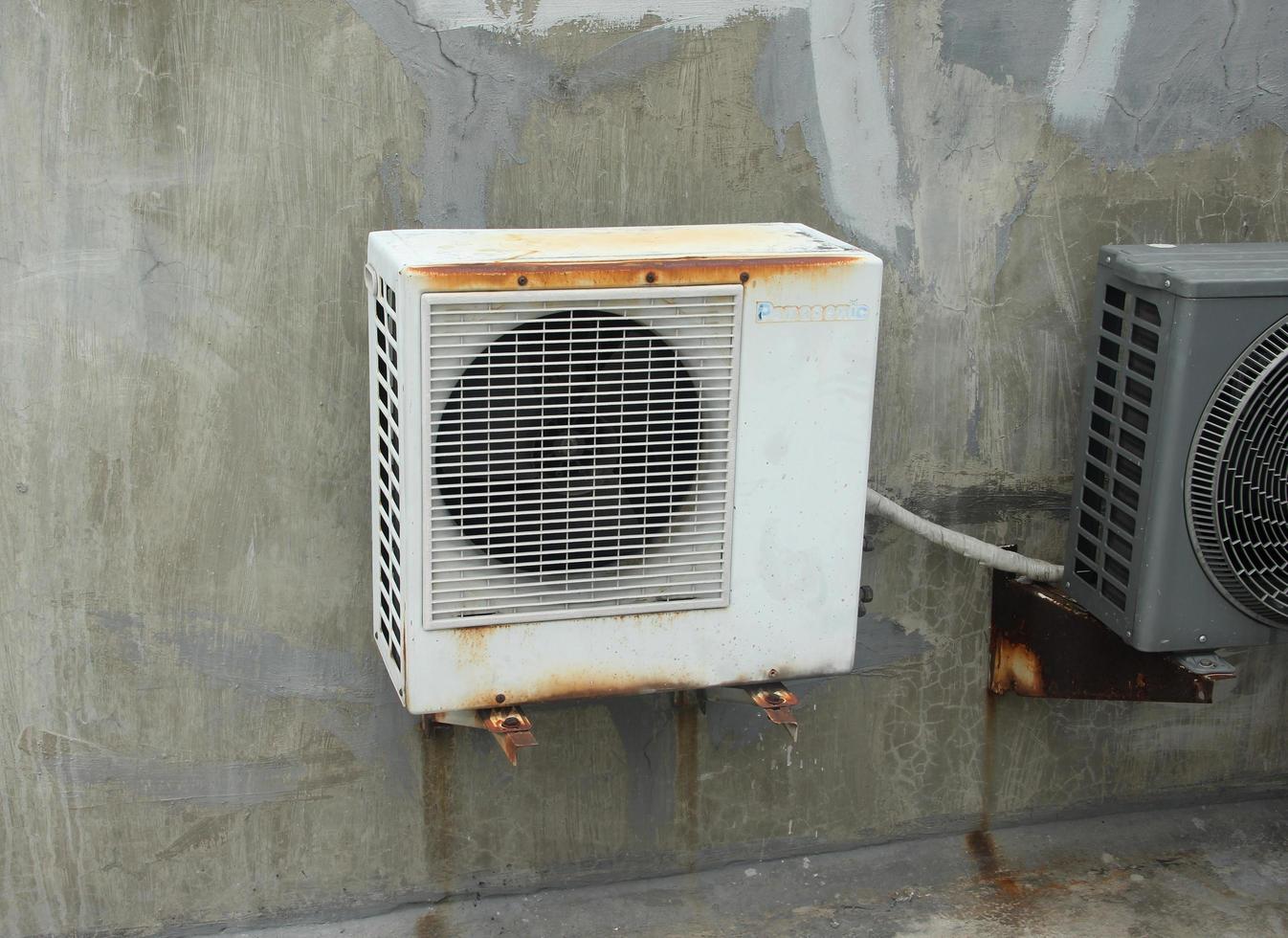 West Jakarta, Indonesia - February 18th, 2023 - White old rusty panasonic ac air conditioner outdoor cooling box isolated on grey cement conrete walls and floors. photo