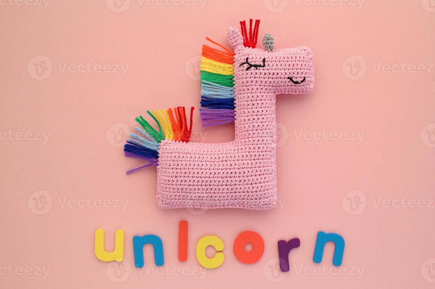 Crochet amigurumi handmade stuffed soft pink unicorn toy with rainbow mane and word on pink background. Handwork hobby. Craft diy newborn pregnancy concept. Knitted doll for little baby. Flat lay photo
