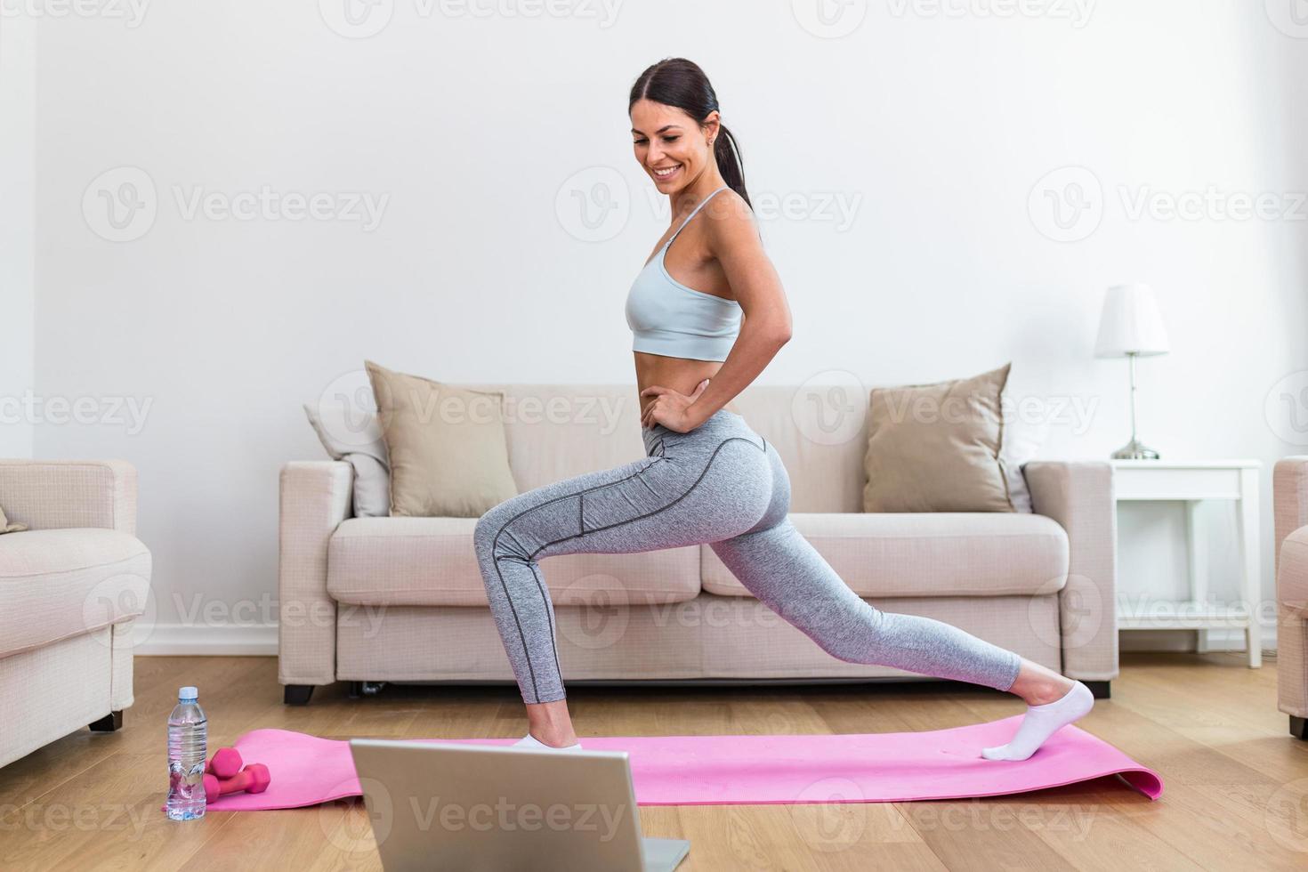 Gorgeous young woman on exercise mat at home and watching workout tutorial online on laptop. Video lesson. Young woman repeating exercises while watching online workout session photo