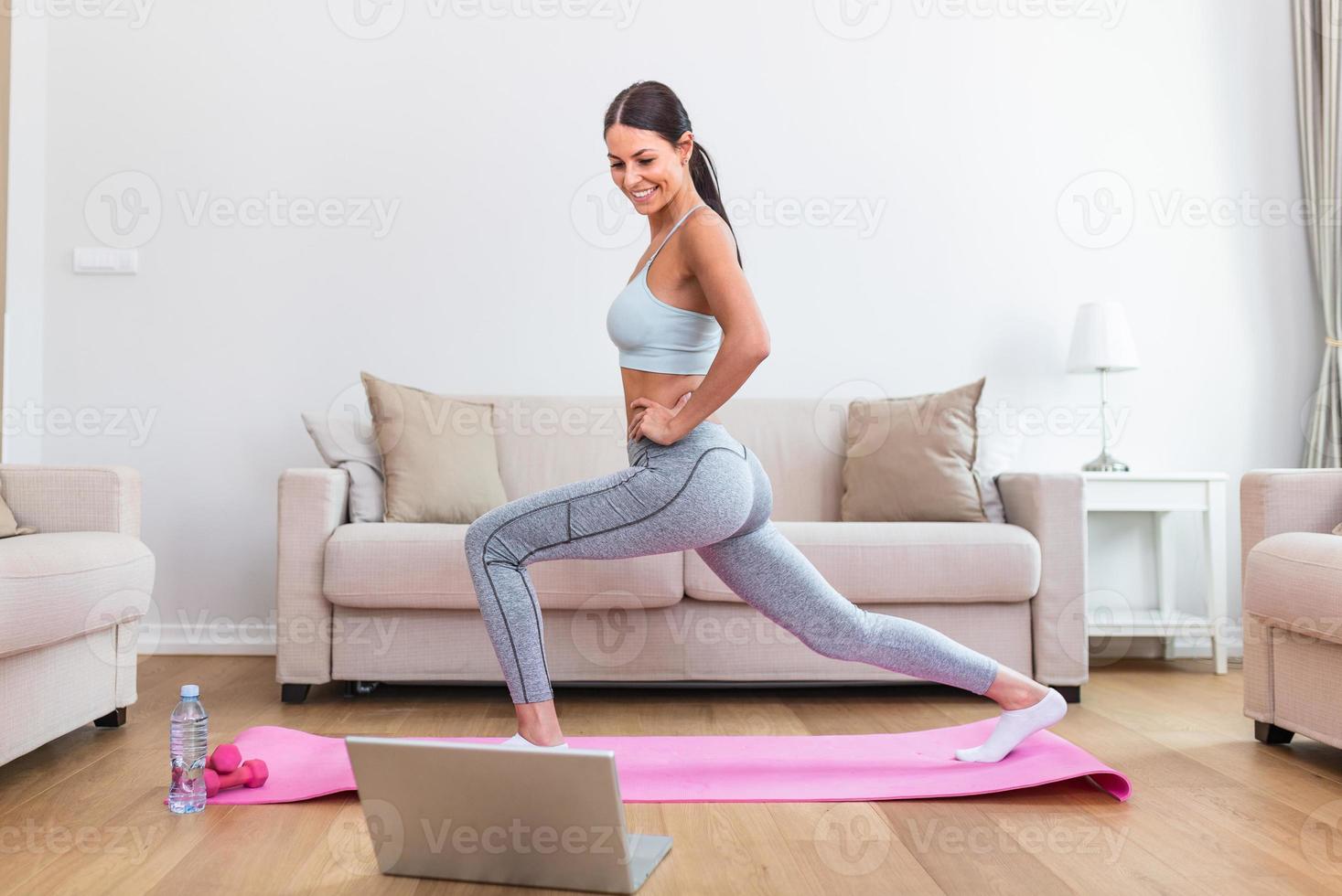 Gorgeous young woman on exercise mat at home and watching workout tutorial online on laptop. Video lesson. Young woman repeating exercises while watching online workout session photo