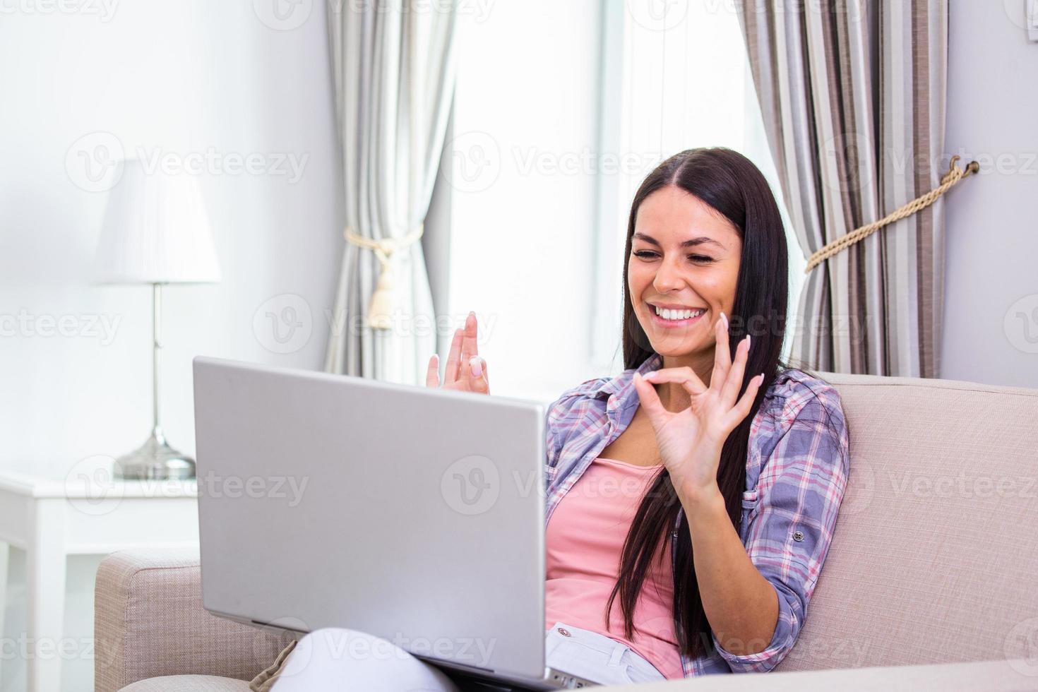 Attractive happy young woman sitting on the sofa using laptop computer, having video chat, waving, talks with her friend about coronavirus and stay home during online video call. Working home photo