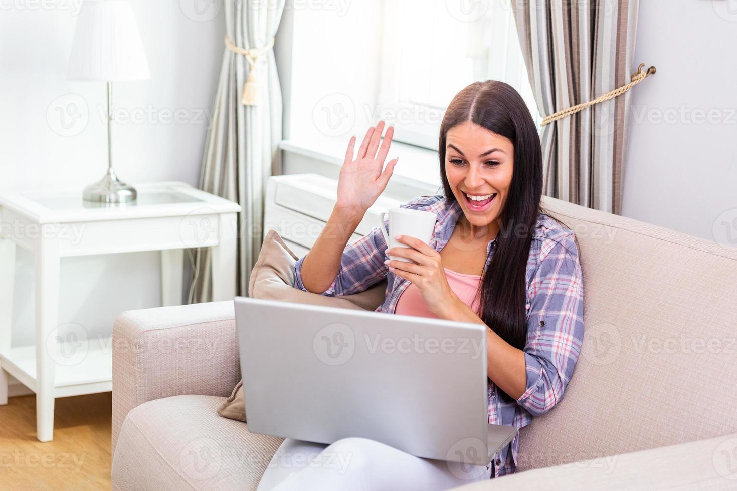 Attractive happy young woman sitting on the sofa using laptop computer, having video chat, waving, talks with her friend about coronavirus and stay home during online video call. Working home photo