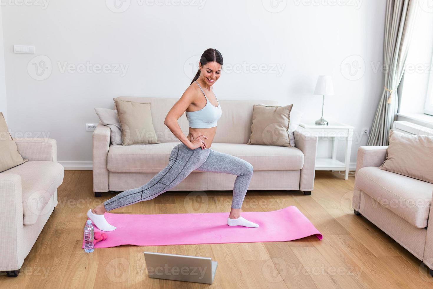 Gorgeous young woman on exercise mat at home and watching workout tutorial online on laptop. Video lesson. Young woman repeating exercises while watching online workout session photo