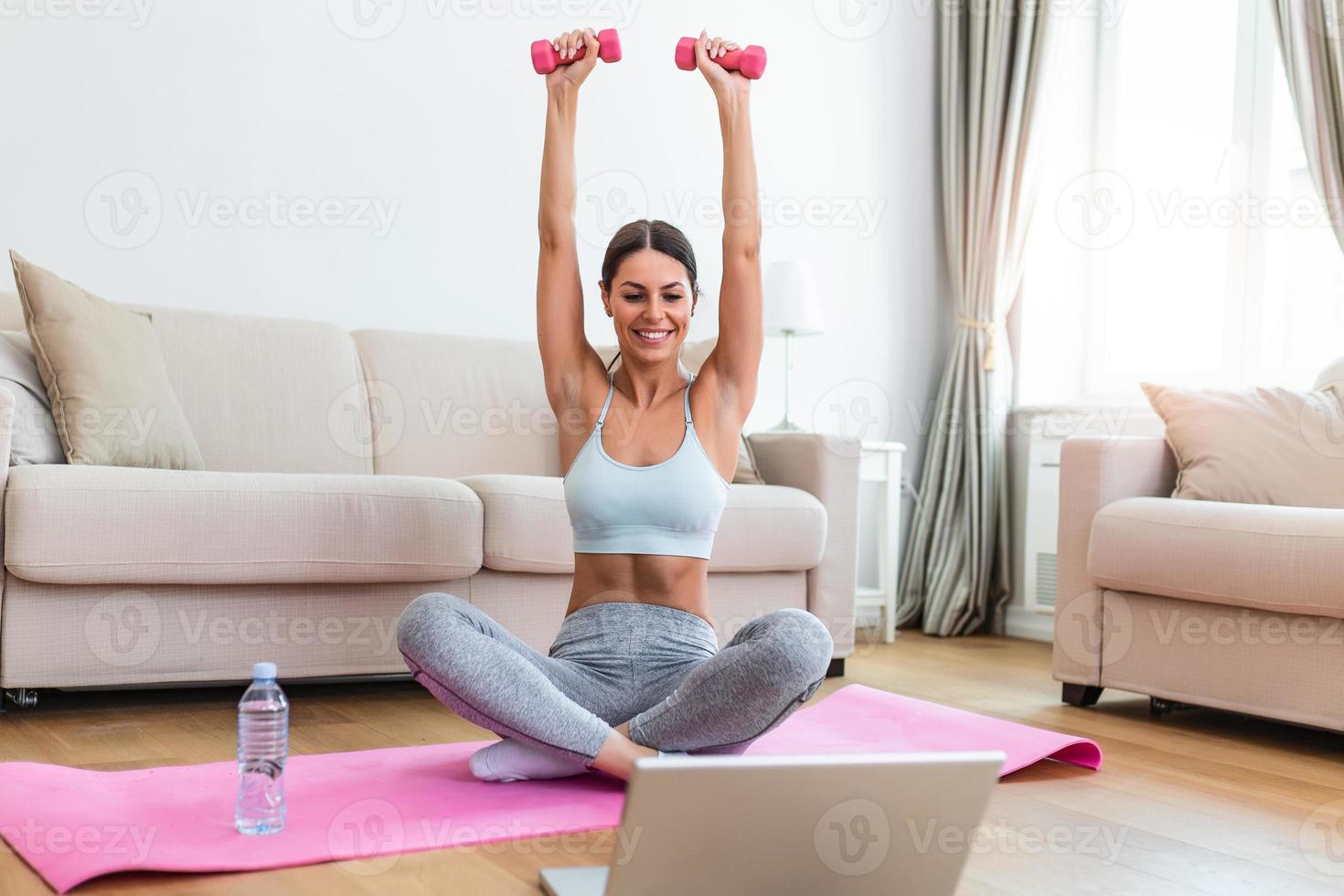 Young woman exercise with dumbbells at home and watching tutorial lesson on laptop computer. happy young female doing arm workout using dumbbells and looking at tutorial on her laptop photo
