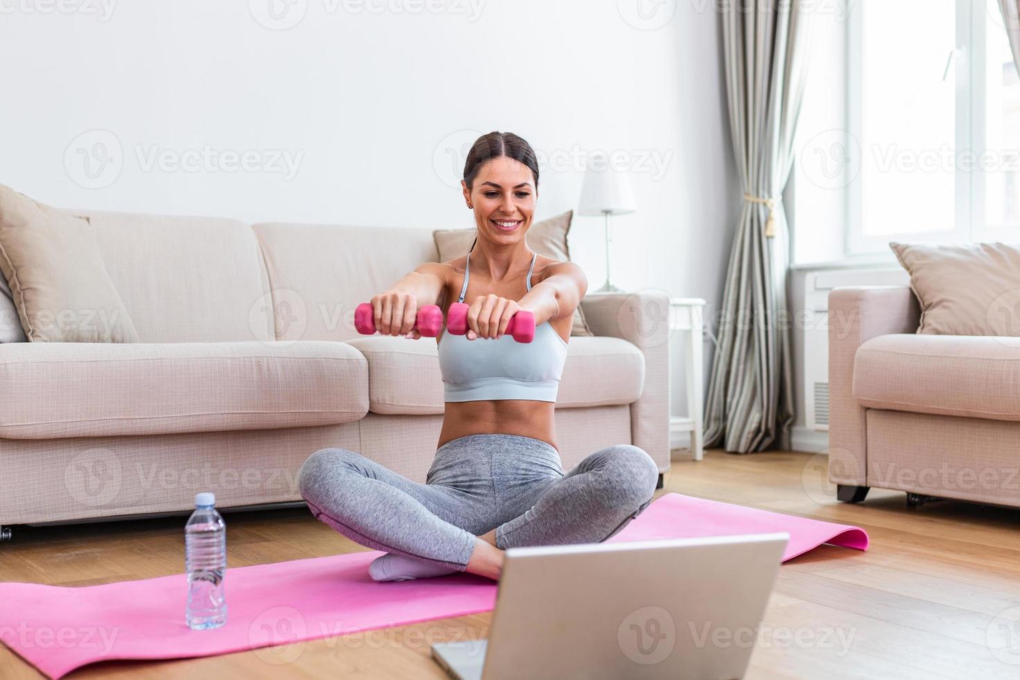 Young woman exercise with dumbbells at home and watching tutorial lesson on laptop computer. happy young female doing arm workout using dumbbells and looking at tutorial on her laptop photo