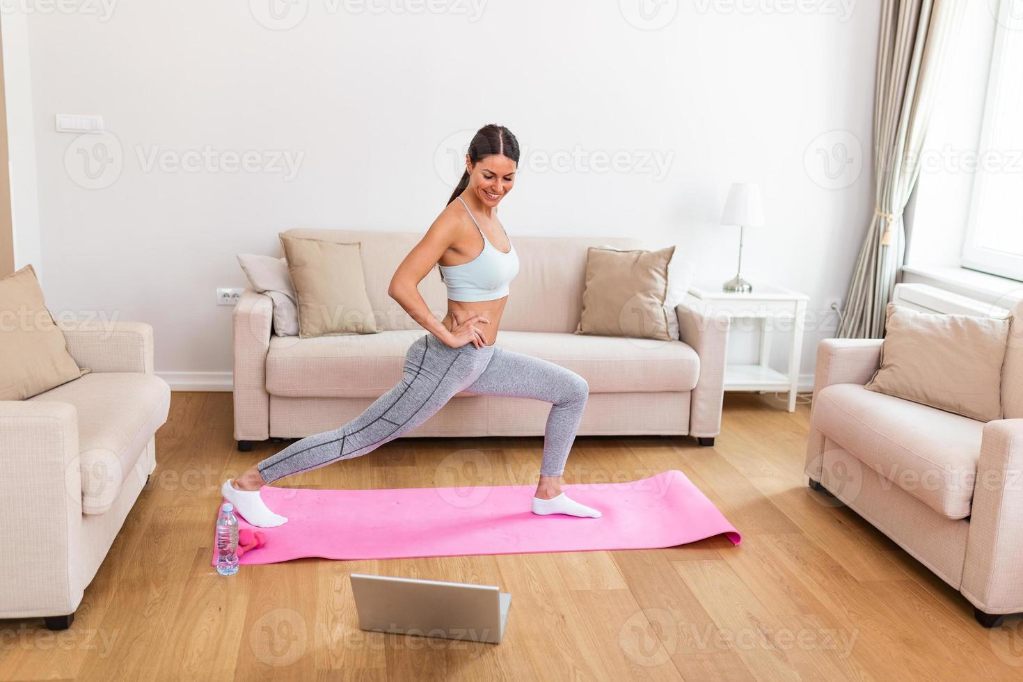 Gorgeous young woman on exercise mat at home and watching workout tutorial online on laptop. Video lesson. Young woman repeating exercises while watching online workout session photo