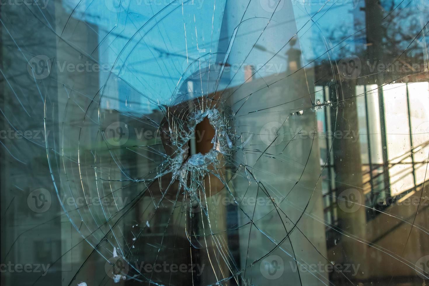 A hole in the window glass with a bullet during a military shooting. Cracks propagate around the hole. Dirty window frame. View of the street from the inside. photo