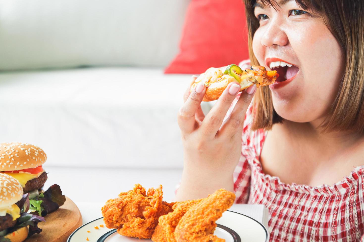 An Asian fat woman is eating pizza and fried chicken on the sofa in her house. The concept of consuming food that causes disease is not good for health photo