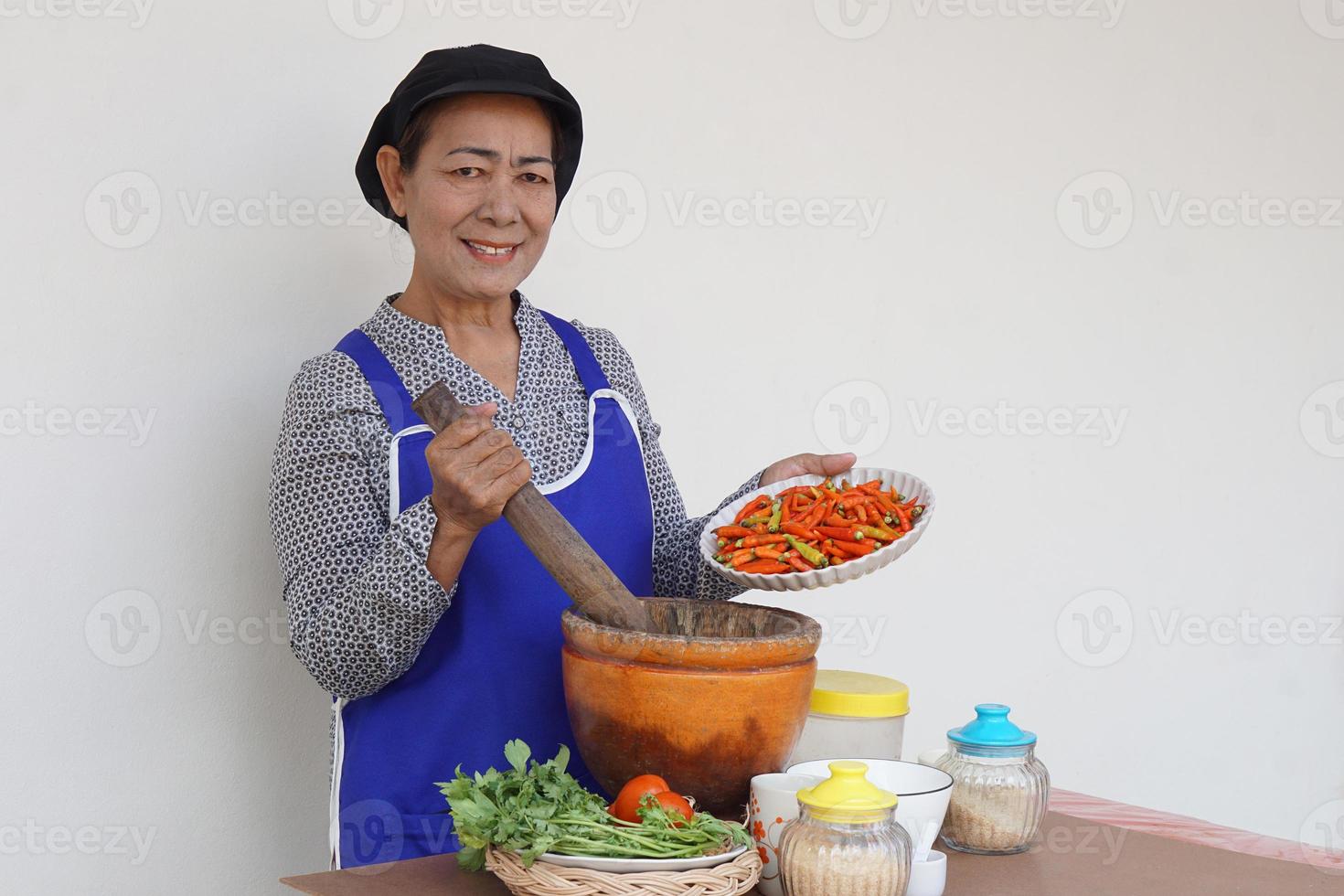 Happy Asian senior woman is cooking, wear chef cap and apron, holds pestle, mortar and plate of chillies. Concept, Cooking for family. Thai kitchen lifestyle. Elderly activity. photo