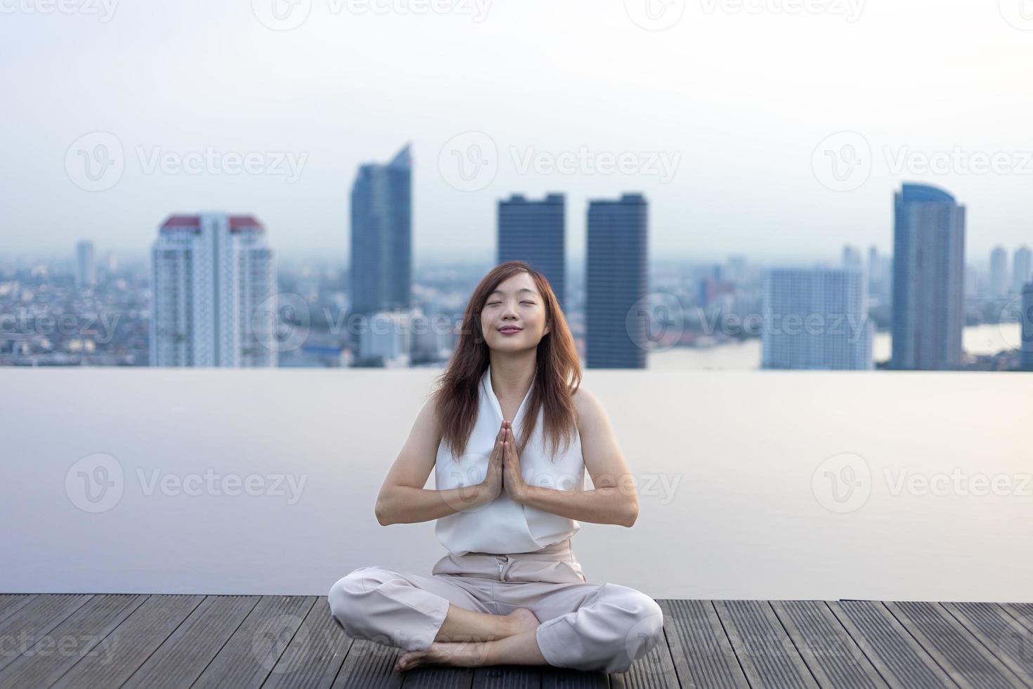 Woman relaxingly practicing meditation at the swimming pool rooftop with the view of urban skyline building to attain happiness from inner peace wisdom concept photo