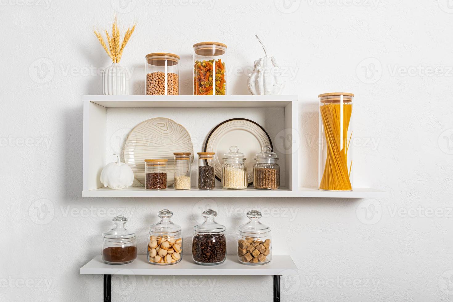 a set of glass and ceramic dishes, glass filled jars with bulk products on white kitchen shelves on textured wall. compact storage. Stylish design. photo