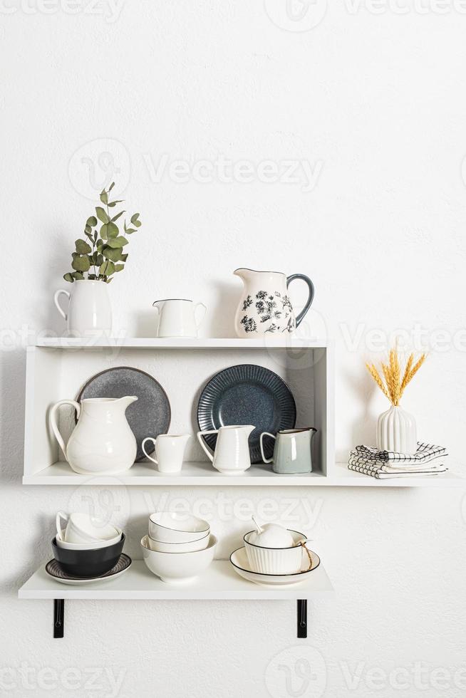 vertical view of the white open kitchen wall shelves with different kinds of jugs, bowls, plates. stylish interior of the kitchen. photo