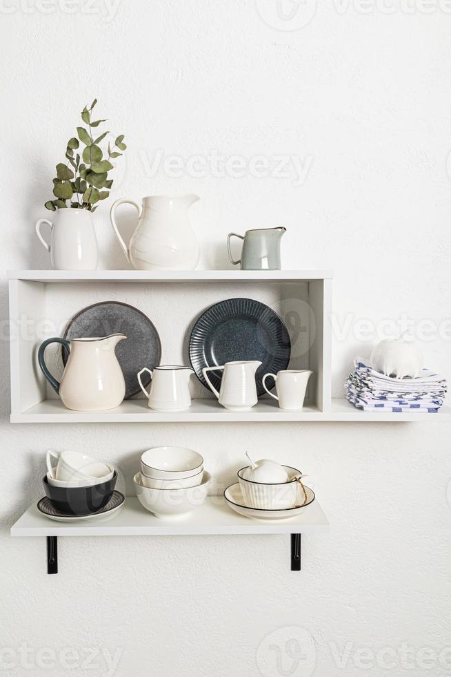 various cookware on open white shelves on a white stylish wall. vertical view. decorative items in the interior of the kitchen. photo
