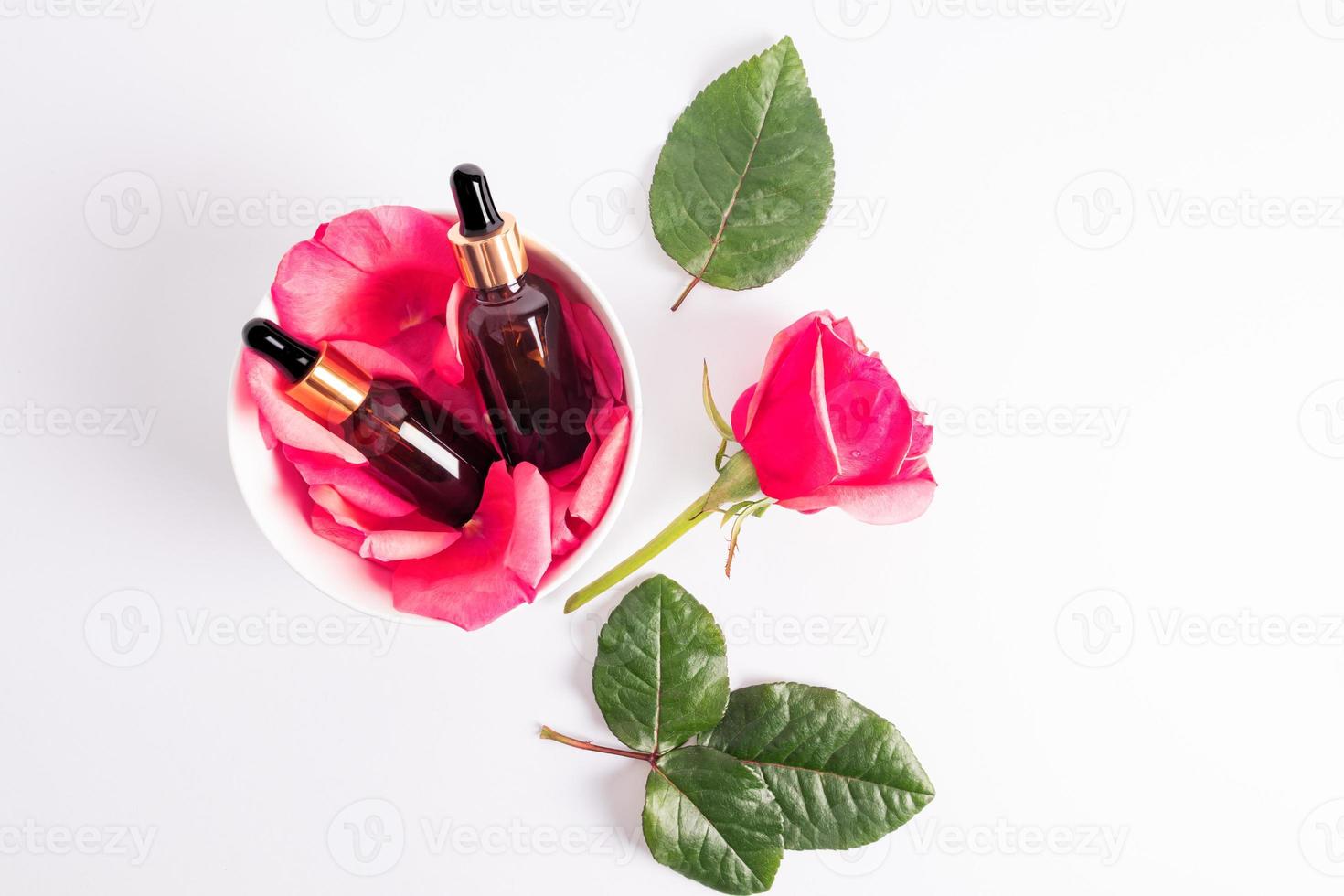 Two cosmetic bottles with a gold cap and a pipette with natural rose oil or moisturizing serum for self-care lie in a bowl with rose petals. Top view. photo