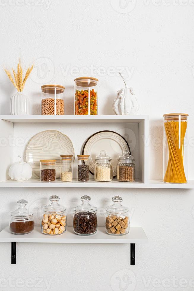 many different glass filled eco-banks for storing bulk products stand on open kitchen shelves against a white textured wall. stylish storage. photo