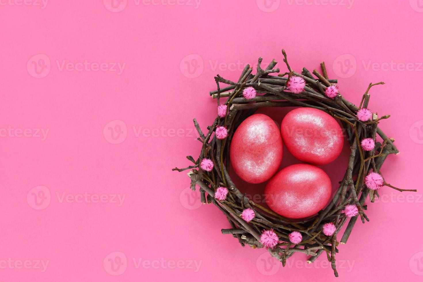 Three 3 Easter eggs colored pink shiny color in nest made of tree branches decorated with pom poms on pink paper background. Glitter paint for eggs. Greeting card for easter holiday. Place for text. photo